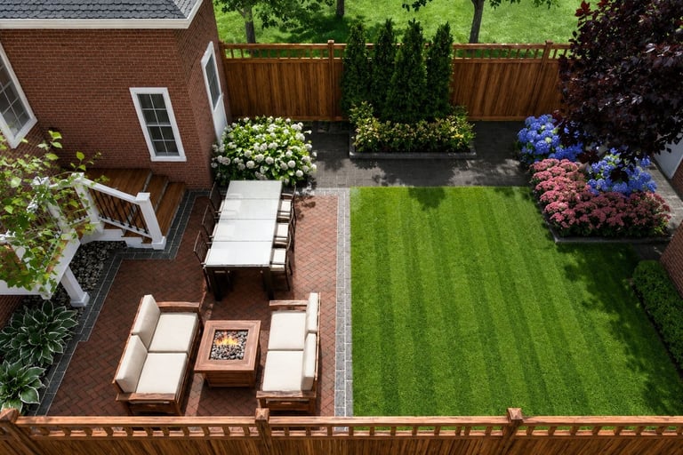Aerial view of a modern backyard landscape with a brick patio, outdoor dining set, fire pit, and lush green lawn.