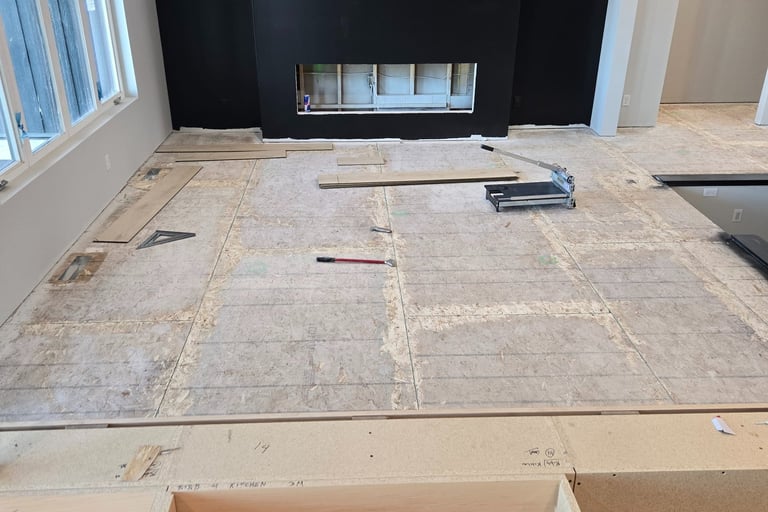 A modern living room under construction featuring a black fireplace wall and subfloor ready for hardwood flooring.