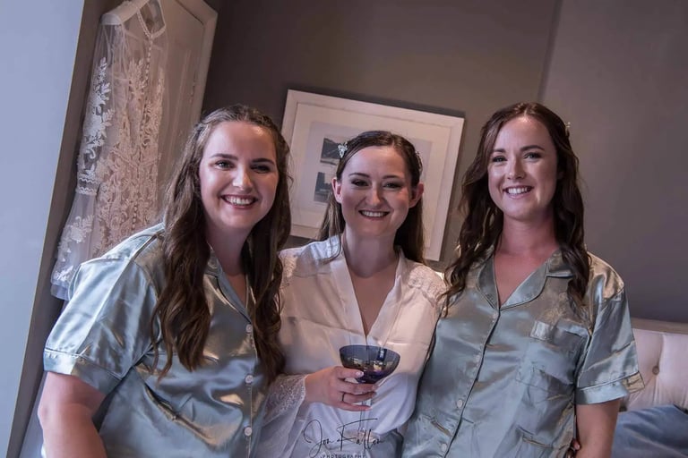 Smiling bride in a white lace robe and bridesmaids in sage green silk pajamas getting ready.