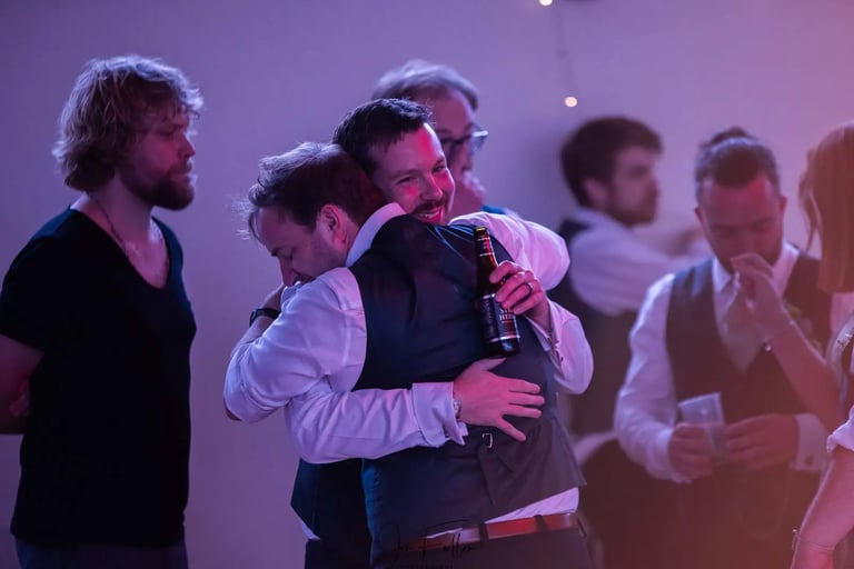Smiling groom hugging a friend at a wedding reception party with guests and drinks in the background.