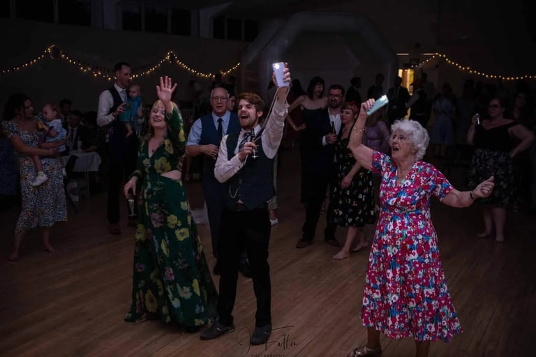 Wedding guests dancing on a wooden floor at a reception with string lights and festive outfits.