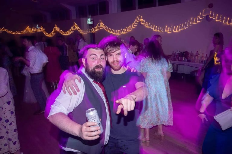 Two friends smiling and dancing under purple event lights at a wedding reception party with string lights.
