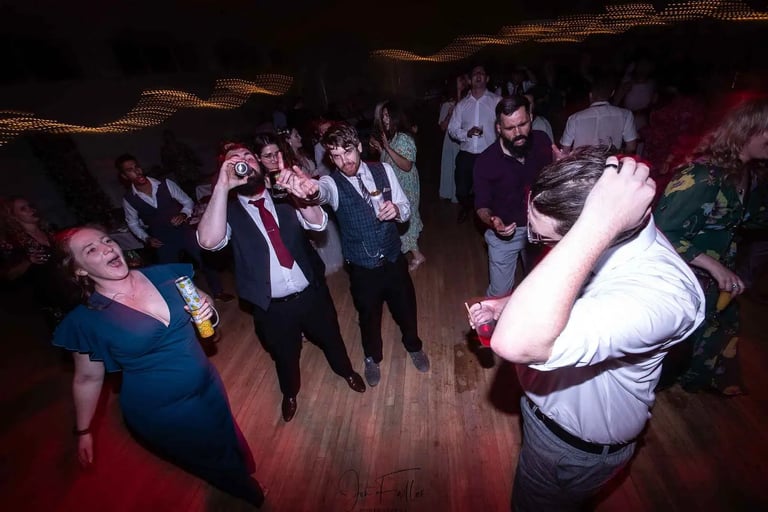 Wedding guests dancing and drinking on a dimly lit dance floor during a lively wedding reception.
