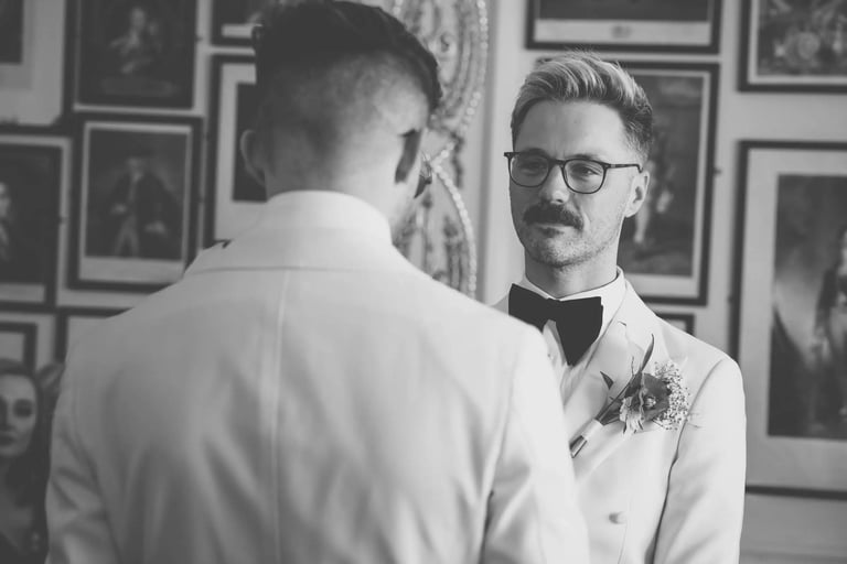 Groom in white tuxedo and bow tie looking at his partner during a wedding ceremony.