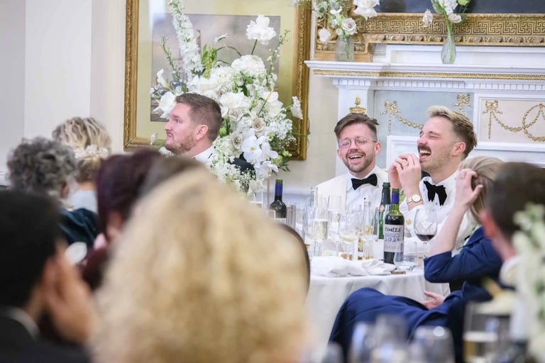 Grooms in white tuxedos laughing at a formal wedding reception dinner table with floral arrangements.