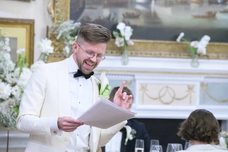 Smiling groom in a white tuxedo giving a wedding speech while holding notes at a reception.