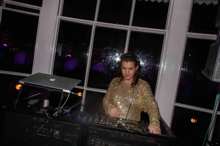 Female DJ in a gold sequin top performing a live set with a mixer and laptop at a night event.