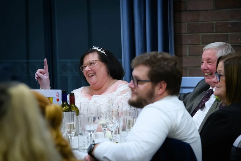 Smiling bride in white lace dress and tiara laughing at a wedding reception dinner table.