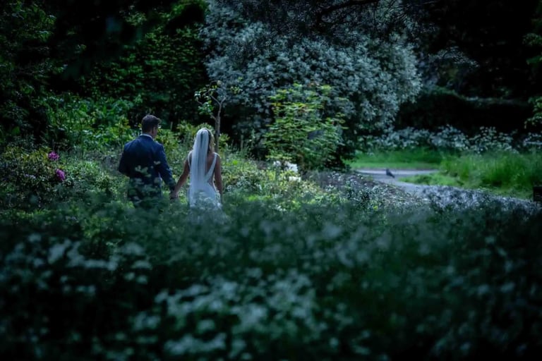 A bride and groom holding hands while walking through a lush green garden at twilight.