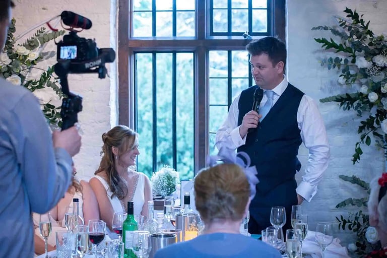Groom giving a wedding speech at the head table being filmed by a professional videographer.