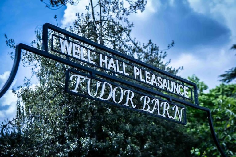 Black wrought iron sign for Well Hall Pleasaunce and Tudor Barn against a blue sky with trees.