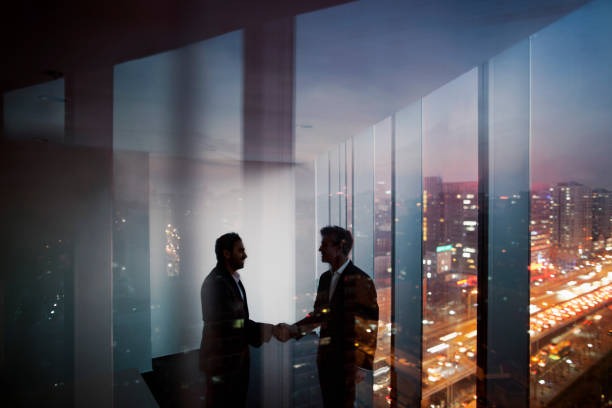 Business partners shaking hands in a high-rise office overlooking a city skyline at sunset.