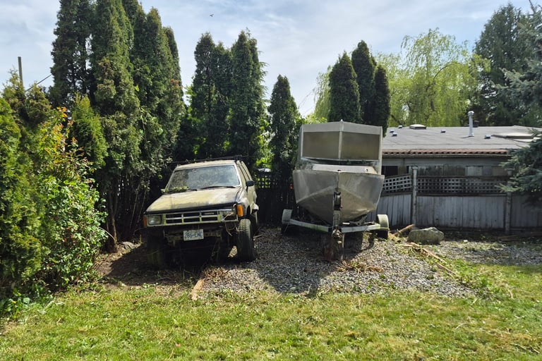 an aluminum boat on a trailer parked beside an older Toyota SUV. Both on a gravel pad in front of an older fence