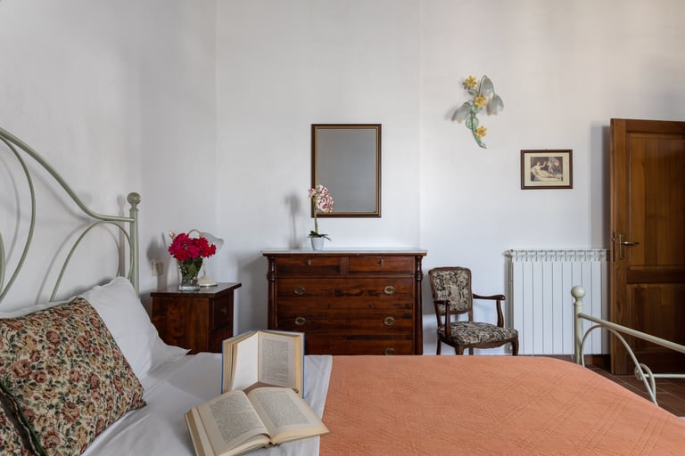 Rustic Italian bedroom interior featuring a wooden dresser, floral pillows, and open books on a bed.