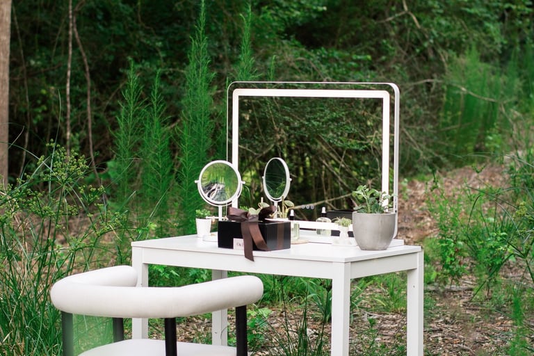 A modern white vanity table with a lighted mirror and armchair set outdoors in a lush green forest.