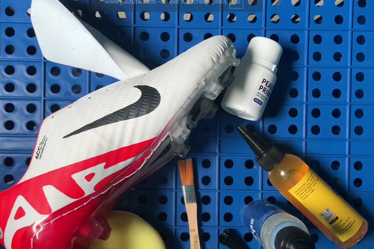 White and red Nike soccer cleat being repaired with shoe glue, primer, and brushes on a blue pegboard.
