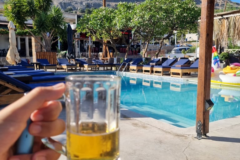 Hand holding a cold beer mug by a sunny resort swimming pool with mountain views.