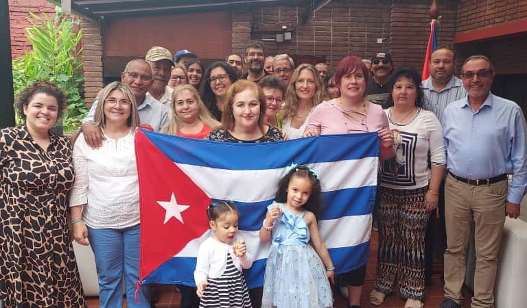 A diverse group of smiling people standing together and holding the national flag of Cuba at an outdoor event.