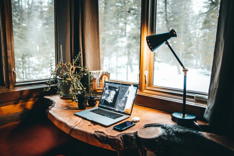 Cozy home office desk with laptop and lamp overlooking a snowy winter forest.