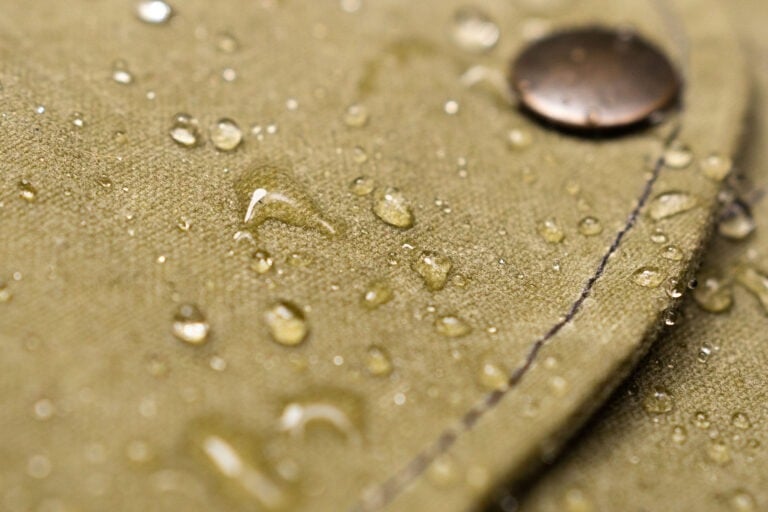 Water droplets beading on a waterproof olive green waxed canvas jacket with a metal button.