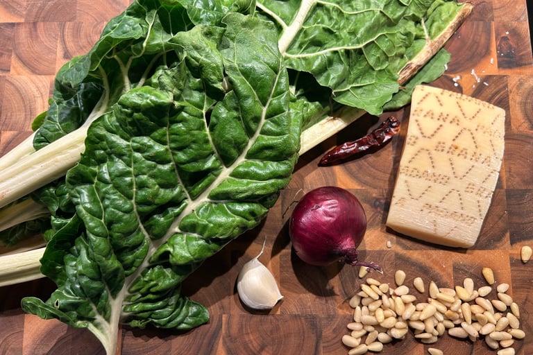 Fresh green chard leaves with a red onion, garlic, pine nuts, and parmesan cheese on a wooden board.
