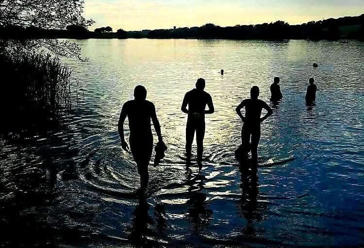 James Oswald Silhouetted swimmers preparing for open water swimming in a calm lake at sunset.