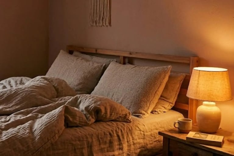 Un letto in legno con biancheria in lino grezzo, comodino in legno con sopra un libro, una tazza e una lampada con luce calda
