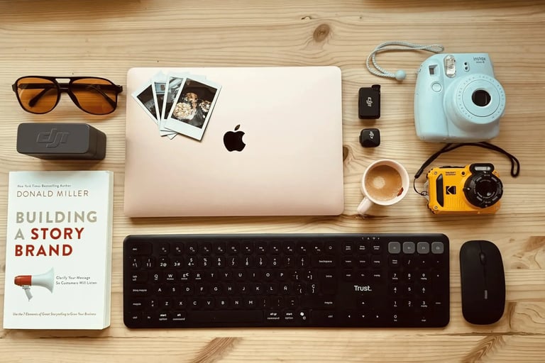 Flat lay of a workspace with a MacBook laptop, Instax camera, Trust keyboard, and Building a Storybrand book.