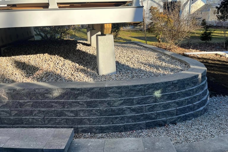 Curved grey block retaining wall supporting a house deck with gravel drainage and concrete piers.