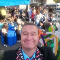 A smiling man takes a selfie at a busy outdoor community festival with balloon animals and crowds.