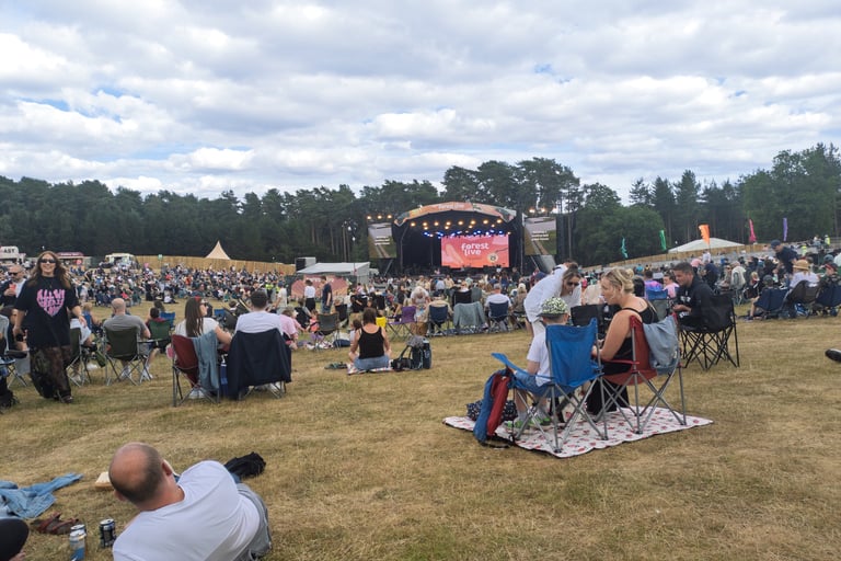 Crowd of people sitting on lawn chairs at an outdoor Forest Live music festival stage in a forest clearing.