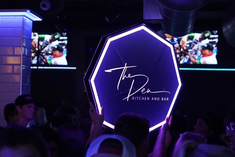 A glowing neon hexagon sign for The Den Kitchen and Bar held up in a crowded nightclub venue.