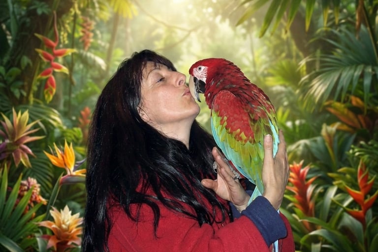A woman kisses a colorful red-and-green macaw parrot in a tropical jungle setting.