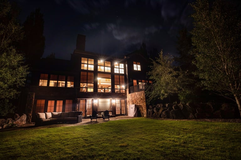 Luxury mountain home with glowing windows at night featuring a stone patio and grassy lawn.