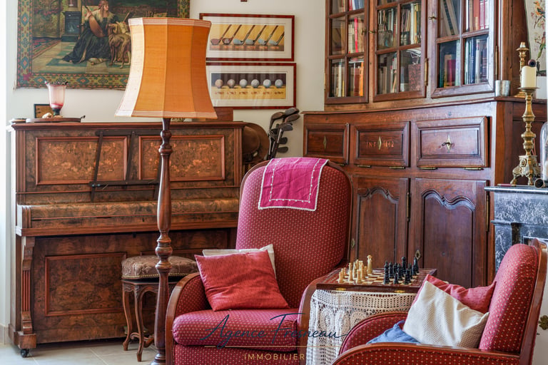 Salon de style vintage avec piano ancien en bois, fauteuils rouges et jeu d’échecs.
