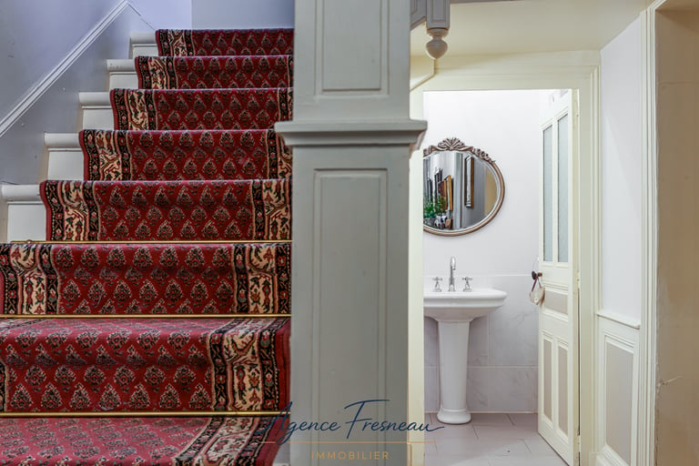 Escalier recouvert d’un tapis rouge à motifs, longeant une salle de bains équipée d’un lavabo.
