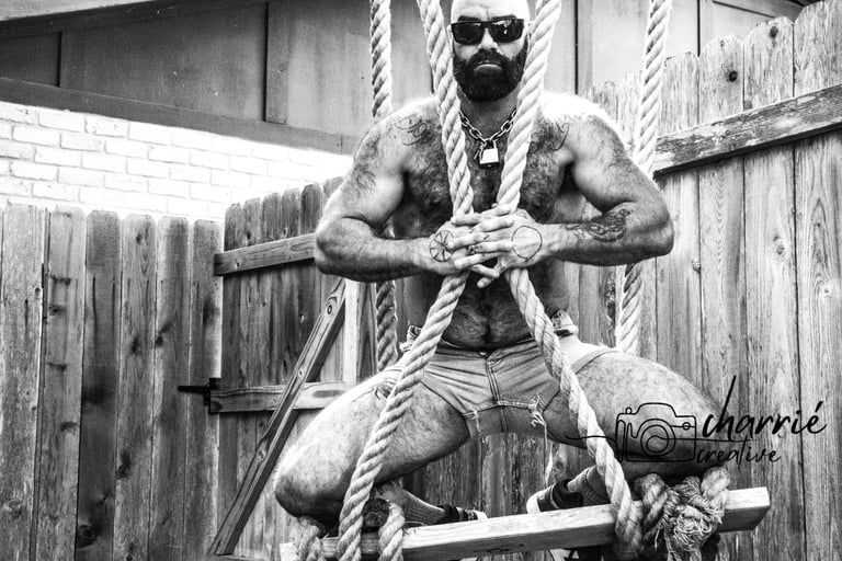 hairy, shirtless bearded man in sunglasses and cutoff shorts hugging ropes