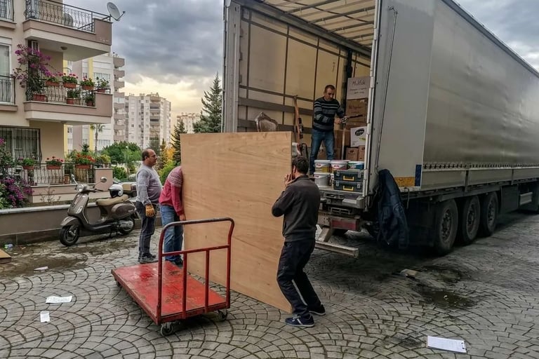Antalya'da bir kamyonun arkasından büyük bir ahşap paneli indiren ve taşıma arabasına yükleyen işçiler.