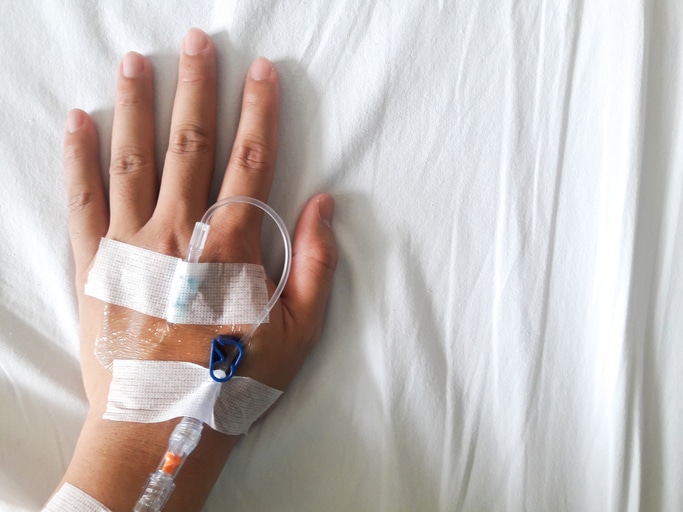 Patient hand with a medical IV drip catheter and bandages on a white hospital bed sheet.