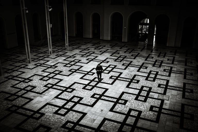 High-contrast black and white photograph of a lone figure framed by dramatic light on a large, patterned stone floor.