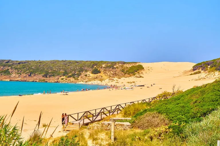 Panoramic view of Bolonia Beach in Tarifa, Spain, featuring sand dunes, turquoise ocean water, and a wooden walkway.