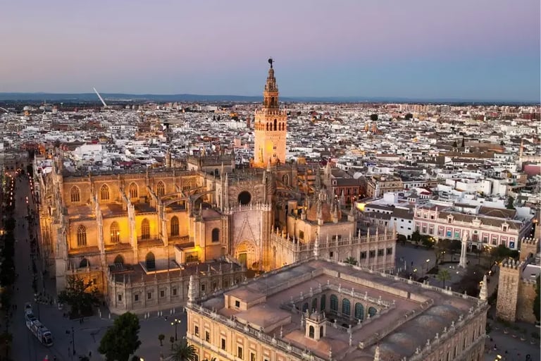 Aerial sunset view of the Seville Cathedral and Giralda tower illuminated against the city skyline in Spain.