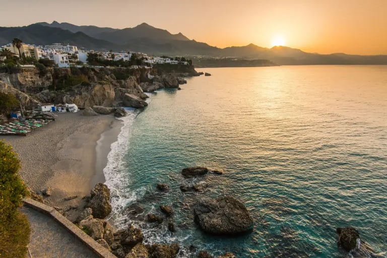 Sunset over the rocky coastline and sandy beach of Nerja, Spain, with mountains in the background.