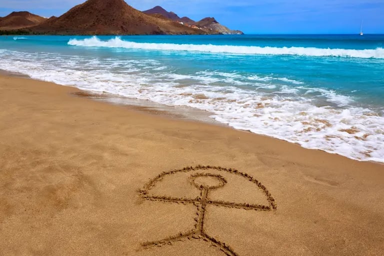 Indalo symbol drawn in the sand on a sunny beach in Almeria, Spain, with ocean waves and mountains.