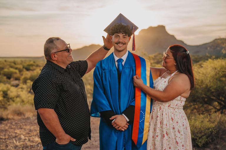 A graduate in a blue gown and cap stands with his parents at sunset in a desert landscape.