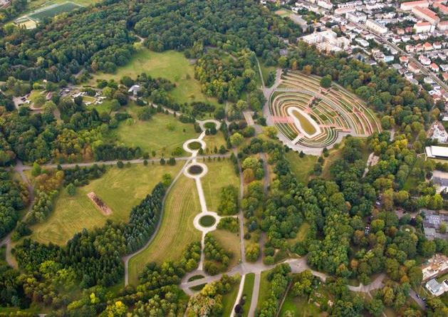 Aerial view of a sprawling city park with lush green trees, circular pathways, and an outdoor amphitheater.