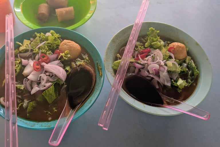 Two bowls of spicy Malaysian laksa soup with noodles, onions, and pink chopsticks on a table.