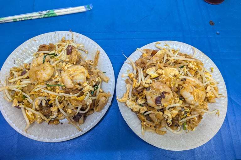 Two plates of authentic Char Kway Teow with prawns and bean sprouts on a blue table.