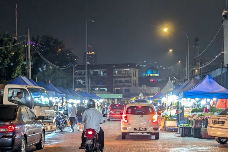 Busy night market street in Malaysia with cars, motorcycles, and blue food stalls under streetlights.