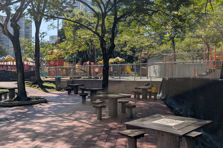 Sunny KLCC park with stone picnic tables and benches near a children's playground and green trees.